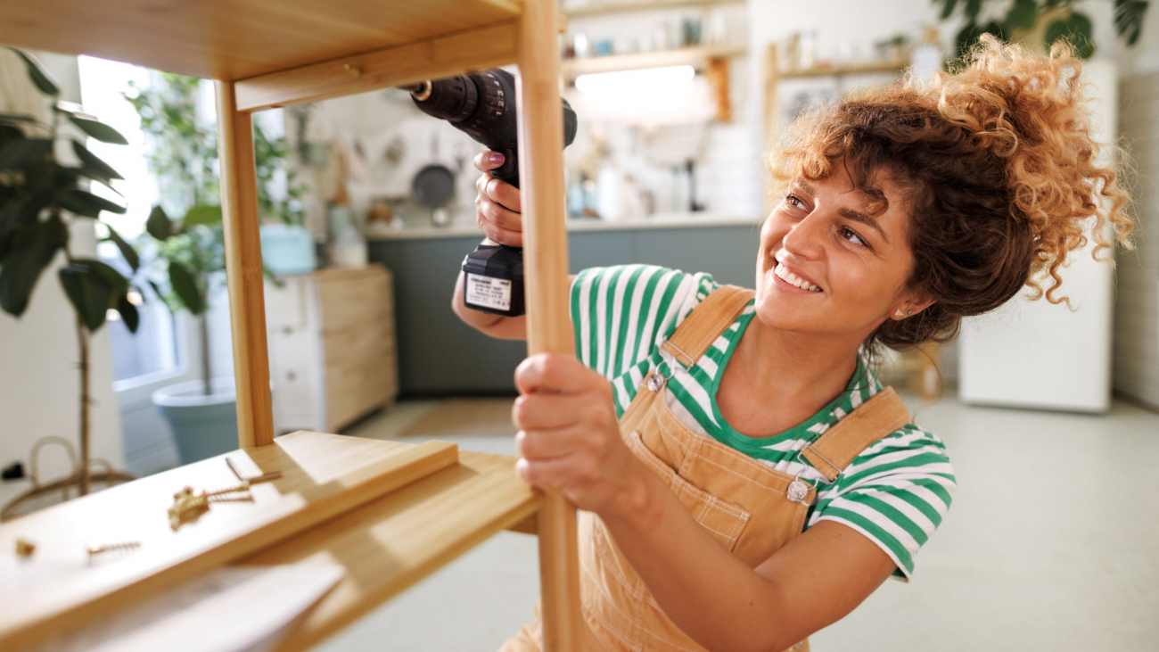 Young beautiful casually clothed woman assembling shelf with electric drill at home